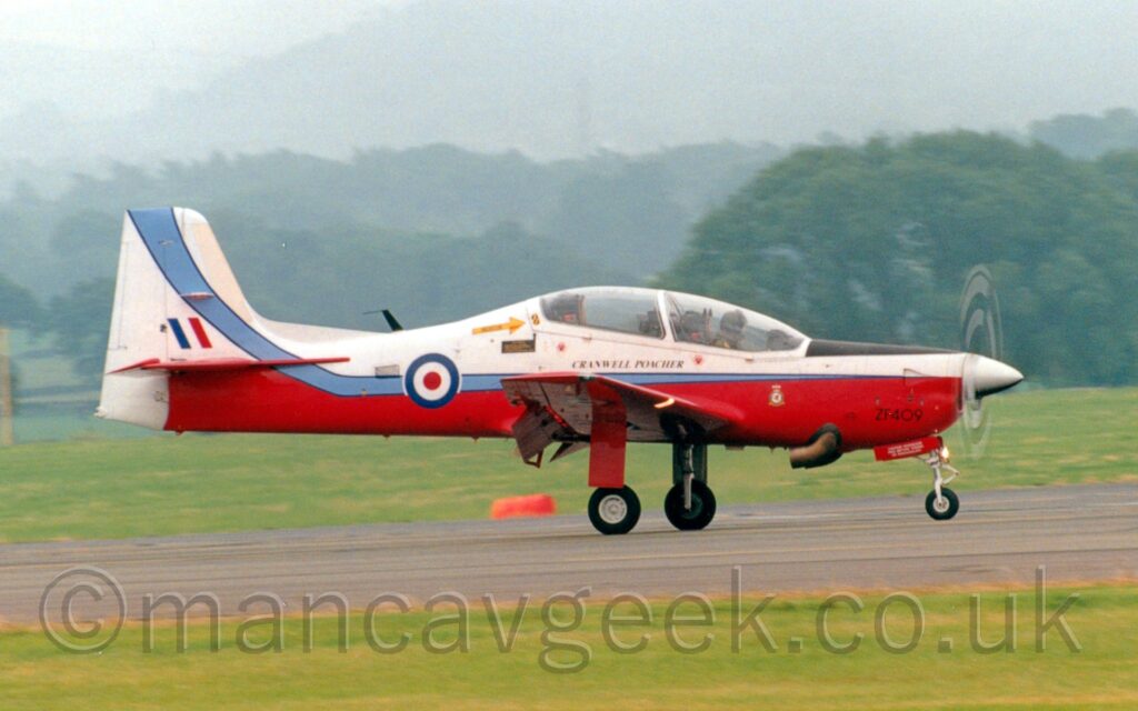 Side view of a white and red, single propellor-engined military training aircraft moving from left to right along a runway. Flaps are extended behind the wings at an extreme angle, suggesting it has just landed. The plane has white upper surfaces, with a red belly and wings, and a pale blue stripe running along the body which sweeps up into the white tail. The black serial "ZF409" is in a non-standard position on the lower front of the engine cowling, with black "Cranwell Poacher" titles under the heavily glazed bubble canopy. There is a red, white, and blue roundel on the rear fuselage, and a blue, white, and red fin flash on the tail. Grass lines the sides of the runway in the foreground, with more grass leading off to trees in the distance, under hazy grey skies.