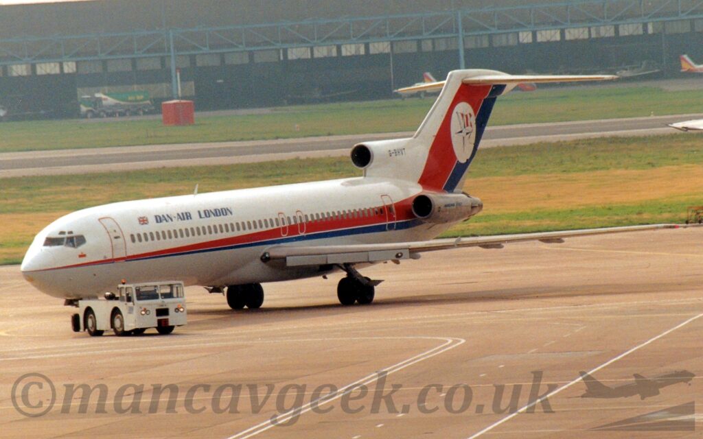 Side view of a 3 engined jet airliner with the engines mounted on the rear fuselage, being towed from right to left, turning back to the right again. The plane is mostly white, with a thin red and blue stripe running along the body, getting wider as it moves rearwards before sweeping up in to the tail. There is blue "Dan-Air London" text on the upper forward fuselage, and the black registration "G-BHVT" on the side of the centre intake above the rear fuselage. There is a white circle on the tail, containing a 4-pointed compass star with a red and white barred flag behind. The plane is being towed by a low-slung white truck turning towards the right of the screen. Grey concrete apron fills the foreground, with grass either side of a runway in the background leading up to a wide black hangar in the distance.