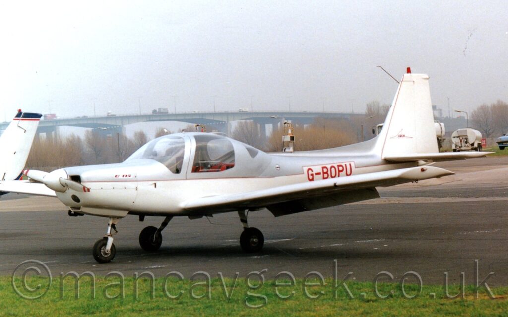 Side view of a white and grey, single engined light aircraft parked on tarmac facing to the left. The plane has grey upper surfaces and white lower half, with red piping separating the 2 sections. The red registration "G-BOPU" is on the rear fuselage. A large bubble canopy covers the 2 seat cockpit. A strip of green grass borders tarmac apron in the foreground, with trees and fuel trucks in the background. A large grey bridge stretches across the frame in the distance, under grey skies.