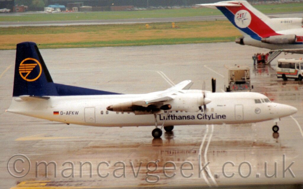 Side view of a white, high-winged, twin propellor-engined airliner taxiing from left to right. The plane has blue "Lufthansa Cityline" titles on the lower forward fuselage, and a dark blue tail with a stylised yellow flying bird in the yellow outline of a circle. Saturated concrete fills most of the screen, it's wet surface reflecting this plane. The rear fuselage, centre engine, and tail of a 3 engined jet airliner parked facing to the right is in the top right corner of the frame, with a number of people climbing the built-in airstairs between the engines as they board the plane. Grass at the top of the frame leads off to black buildings in the distance.
