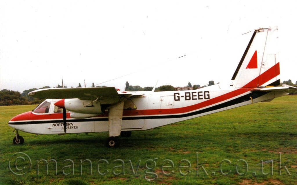 Side view of a small, boxy, white, high-winged, twin propellor-engined airliner parked on grass facing to the left. The plane is mostly white, with a thick red and thin black stripe running along the body before swooping up into the lower reaches of the tail. There are small, black "Northern Airlines" titles on the lower forward fuselage, and larger, black registration "G-BEEG" on the upper rear. The landing gear is fixed, with a nose-wheel at the front, and the main wheels on long white struts that come down from the wings, just inboard of the engines. Green grass fills the foreground, leading up to trees lining the airfield perimeter in the distance, under flat grey skies.