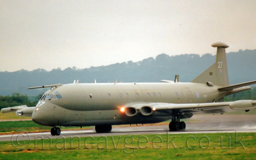 Side view of a 4 jet-engined military patrol aircraft moving at high speed from right to left along a runway, with flaps extended from behind the wings. The plane has yellowy-grey upper surfaces, and lower surfaces in a slightly lighter shade, and a pale grey belly, with large white numbers "27" on the nose and tail. There is a small, pastel blue and red roundel on the rear fuselage, next to the white serial "XV227", and a small, pastel red and blue fin flash on the tail. A bright light is shining in the wing root, on one side of the engine air intakes built in to the wing, with another, smaller, light on the other side of the intakes. A fixed air-to-air refuelling probe extends forwards from the top of the cockpit. Lush green grass fills the foreground, with trees on a hill in the background slowly vanishing in to haze.