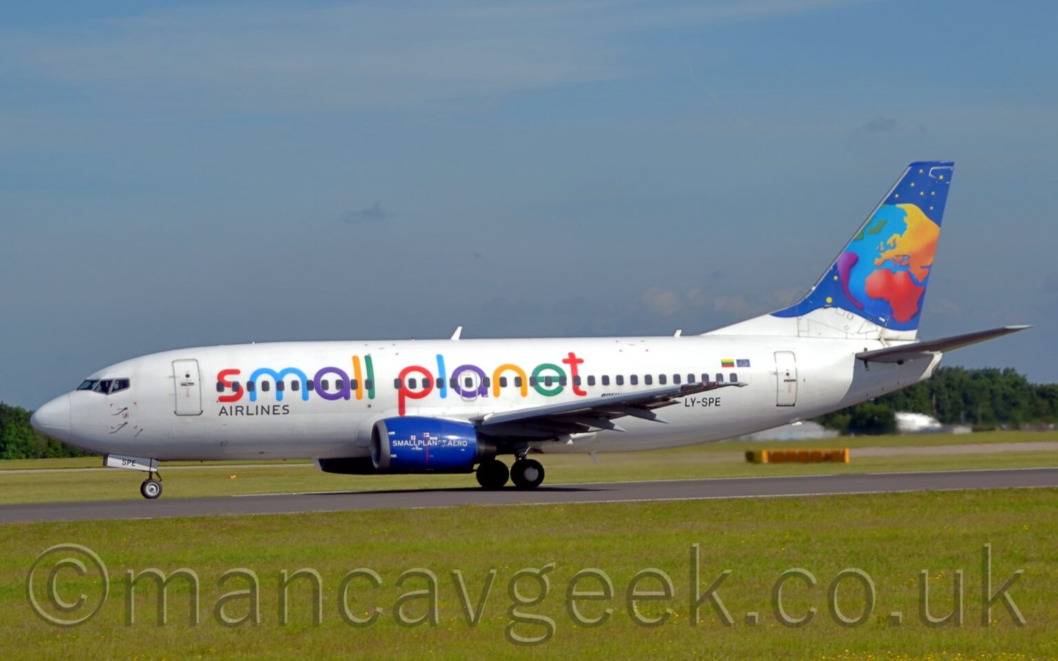 Side view of a twin engined jet airliner accelerating from right to left along a black runway. The plane is mostly white, with multi-coloured "Small Planet" titles along the fuselage, successive letters in red, blue, purple, yellow, and green. The black registration "LY-SPE" is on the lower rear fuselage. The engine pods mounted under the wings are a deep blue, with white "SmallPlanet.Aero" text. The tail is dark blue, with a stylised multi-coloured globe and yellow stars. Lush green grass fills the foreground, with tall green trees in the distance behind the plane on either side, under blue sky with smears of cloud.