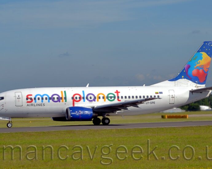 Side view of a twin engined jet airliner accelerating from right to left along a black runway. The plane is mostly white, with multi-coloured "Small Planet" titles along the fuselage, successive letters in red, blue, purple, yellow, and green. The black registration "LY-SPE" is on the lower rear fuselage. The engine pods mounted under the wings are a deep blue, with white "SmallPlanet.Aero" text. The tail is dark blue, with a stylised multi-coloured globe and yellow stars. Lush green grass fills the foreground, with tall green trees in the distance behind the plane on either side, under blue sky with smears of cloud.