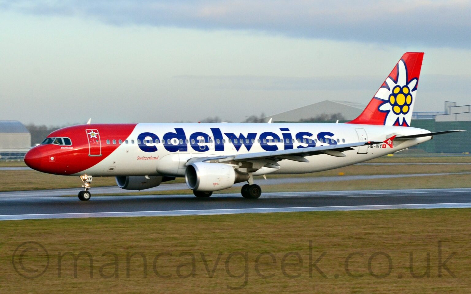 Side view of a twin engined jet airliner accelerating from right to left along a black runway. The plane is largely white, with a diagonal red band wrapped around the nose and forward fuselage, and a stylised white flower with a yellow centre on the forward cabin door. There are large, billboard-style "Edelweiss" titles on the upper fuselage, with smaller, red "Switzerland" titles in an italic font just ahead of the wing box, and the black registration "HB-IHY" on the lower rear fuselage, next to a small Swiss flag. The red tail has a larger version of the white and yellow flower logo, repeated in a smaller form on the vertical wingtip fences. Green grass lines the black runway in the foreground, with taxiways and more grass in the background, with grey hangars in the distance on either side of the image, and pale blue sky with smears of grey cloud filling the rest of the frame.