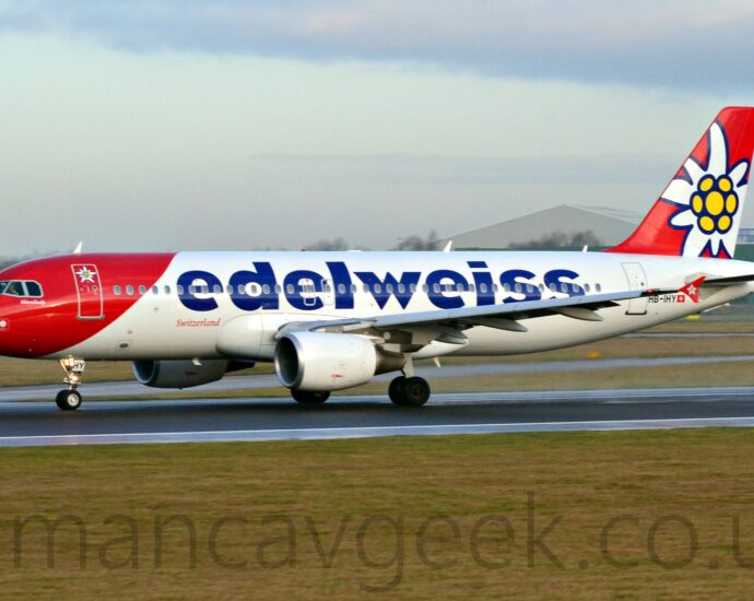 Side view of a twin engined jet airliner accelerating from right to left along a black runway. The plane is largely white, with a diagonal red band wrapped around the nose and forward fuselage, and a stylised white flower with a yellow centre on the forward cabin door. There are large, billboard-style "Edelweiss" titles on the upper fuselage, with smaller, red "Switzerland" titles in an italic font just ahead of the wing box, and the black registration "HB-IHY" on the lower rear fuselage, next to a small Swiss flag. The red tail has a larger version of the white and yellow flower logo, repeated in a smaller form on the vertical wingtip fences. Green grass lines the black runway in the foreground, with taxiways and more grass in the background, with grey hangars in the distance on either side of the image, and pale blue sky with smears of grey cloud filling the rest of the frame.