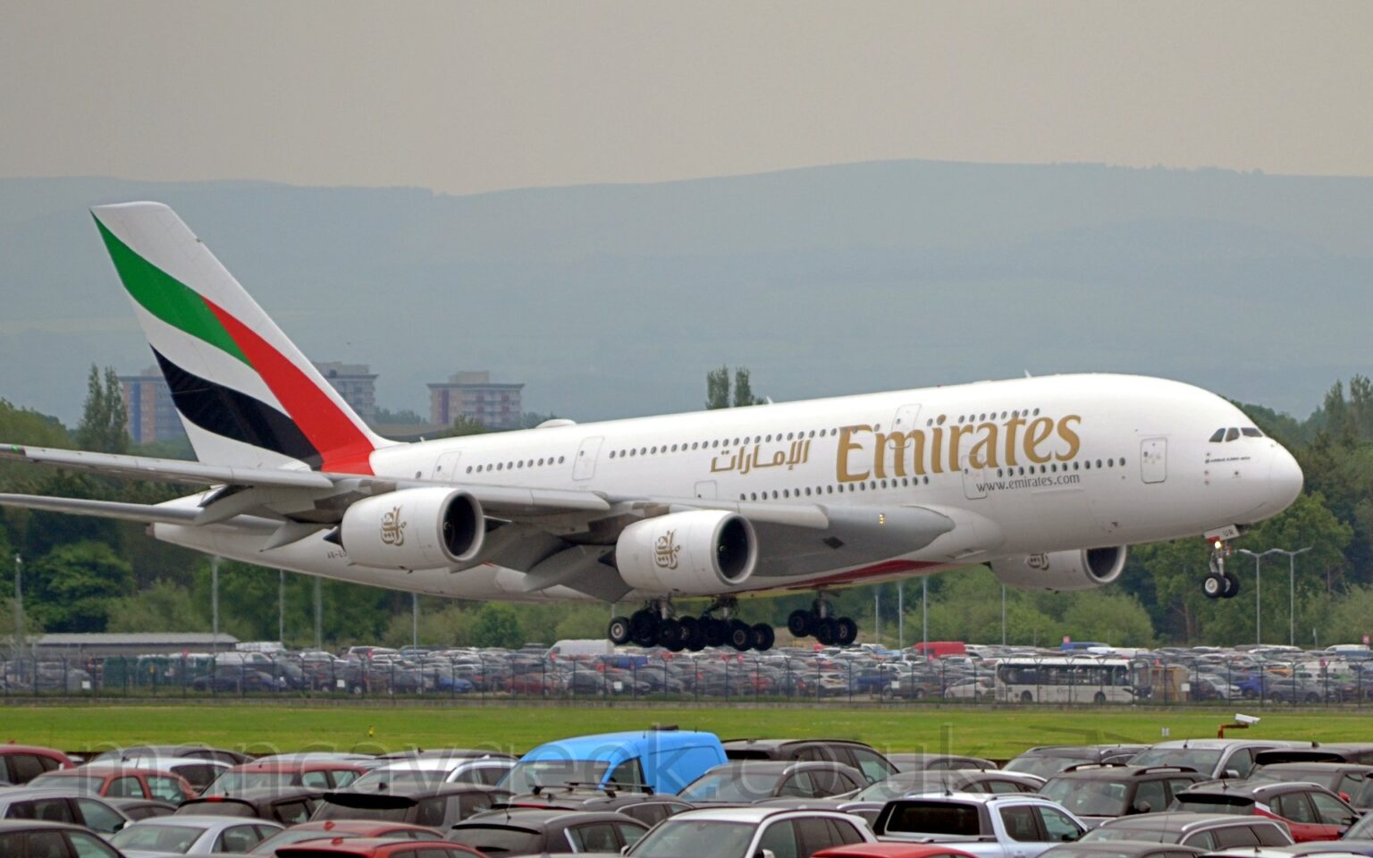 Side view of a very large, white, 4 engined jet airliner flying from left to right at a very low altitude, with undercarriage extended and flaps deployed from behind the wing, suggesting it is just about to land. There are large golden "Emirates" titles on the forward fuselage, repeated in a smaller Arabic script over the wings. The web address "www.emirates.com" is on the lower forward fuselage, also in gold. There is a golden logo on each engine pod, made up of the Arabic letters that make up the word that trans-literates as "Etihad", but re-arranged in a decorative pattern. The tail has the flag of the United Arab Emirates (red vertical bar on the right, with green, white, and black horizontal stripes on the left), looking as if it is waving in the breeze. In the foreground, a mass of parked cars, mostly black but with a few scattered reds, and a small blue van in the middle, are packed densely along the bottom of the frame, with a strip of green grass leading up to more parked cars on the far side of the runway, behind a black chainlink fence. Trees beyond that lead up to hills shrouded in mist in the distance, under grey skies.