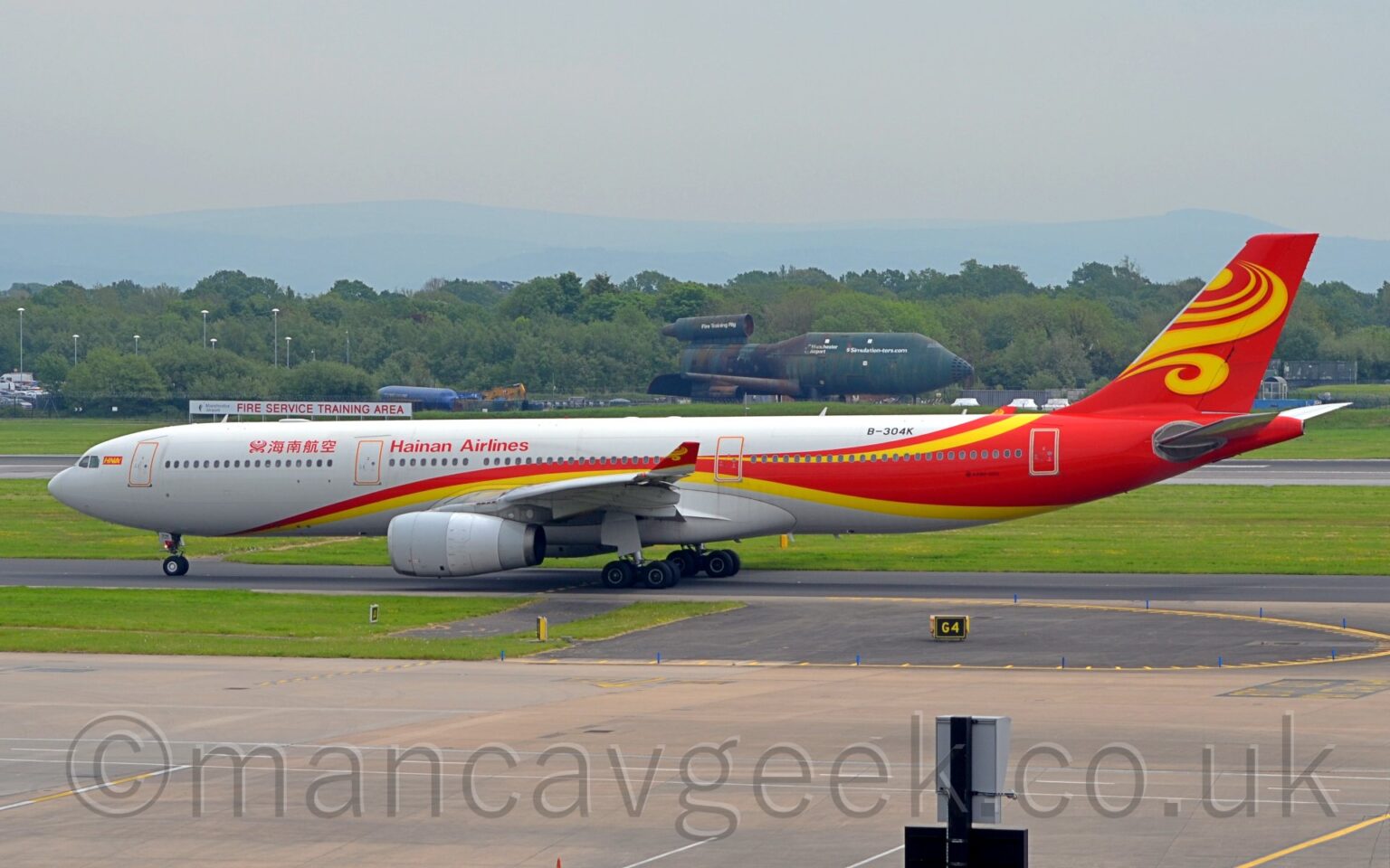 Side view of a twin engined jet airliner taxiing from right to left. The plane is mostly white, with a red and yellow stripe swooping from under the lower forward fuselage and over the wing, with the yellow descending back under the rear fuselage, while the red takes over the rear fuselage and tail, with additional yellow swirls at the top and on the tail. There are red "Hainan Airlines" titles on the upper forward fuselage, in a Chinese script forward if the mid-cabin door, and in English aft of that door. The black registration "B-304K" is on the upper rear fuselage. Grey concrete apron and black tarmac taxiway fill the foreground, with trees stretching across the background under hazy grey sky. In the middle of the background, a green fire training rig, vaguely resembling a 3-engined Boeing 747, is sitting there looking a bit sorry for itself.
