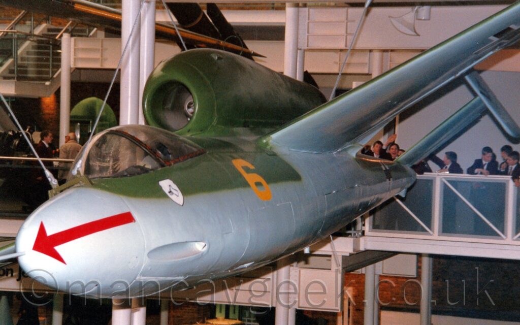 Front view of a green and grey, single-engined World War 2-era military jet fighter in a museum, hanging from the ceiling as if it is flying from right to left and slightly downwards. It's upper surfaces are green, with grey lower surfaces, with a yellow number "6" on the forward fuselage just behind the cockpit, and a red arrow pointing forward on the grey nosecone. The engine is mounted on top of the fuselage, above and behind the cockpit. There are twin tailplanes, canted inwards, mounted on the tips of the horizontal stabiliser. Crowds of schoolboys wearing white shirts and mauve blazers are gathered on a balcony behind the plane, one of them is waving at the camera.