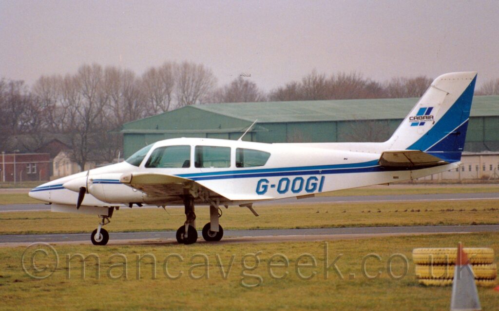 Side view of a twin propellor-engined light aircraft, looking rather tail heavy with it's long, spindly undercarriage so far forward. The plane is mostly white, with light and dark blue stripes running along the body, before turning up into the lower reaches of the tail. The blue registration "G-OOGI" is on the lower rear fuselage, with a slight grey drop shadow. A pair of short, vertical stripes is on the tail, overlaid with the black text "CabAir Flying School". Green grass fills the foreground, with a grey taxiway and more grass in the background leading up to a green hangar in the distance, with bare trees behind that, under grim grey sky.