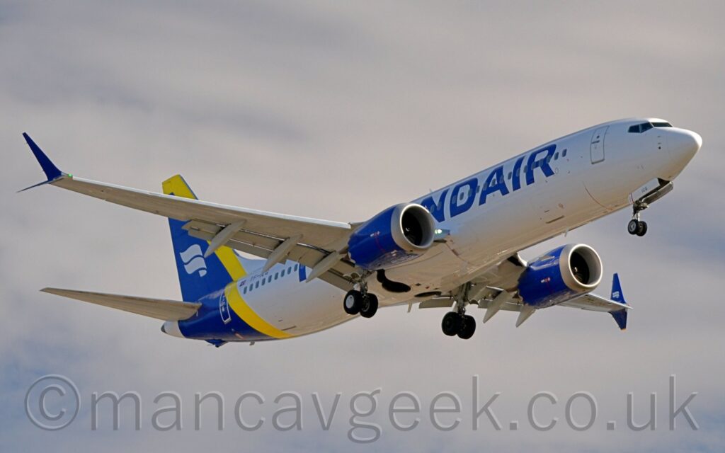 On This Day 6th March 2025. TF-ICG, Boeing 737-8 MAX, IcelandAir, on final approach to Runway 23 Right at Manchester Airport, 6th March 2025, as seen from ShadowMoss Road. #Manchester #MAN #EGCC #Runway23Right #ShadowMossRoad #Boeing #B737 #Max #IcelandAir #AvGeek #aviation #planespotting #photography Manchester, MAN, EGCC, Runway23Right, ShadsowMossRoad, Boeing, B737, Max, IcelandAir Low side view of a twin engined jet airliner flying from left to right at a very low altitude, with undercarriage lowered and flaps extended from behind the wings, suggesting it is about to land. The plane is mostly white, with billboard-style blue "IcelandAir" titles on the mid-fuselage, mostly over the wings. The tail and rear fuselage is blue, fronted with a wide yellow stripe, and a pair of white waving ribbons in the middle. The waving ribbons are also on the inside faces of the blue upturned wingtips, the outer faces being plain blue. The engine pods, mounted under the wings, are the same dark blue. Cloudy blue sky with hints of blue fill the rest of the frame.