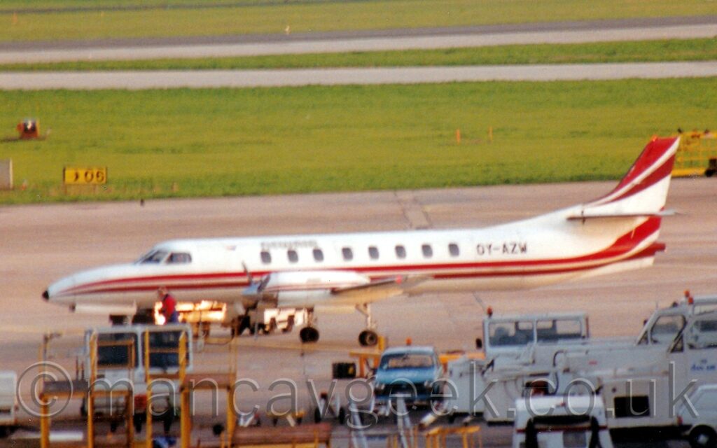 Slightly blurry side view of a long, thin, twin propellor-engined parked facing to the left. The plane is mostly white, with a pair of red stripes running along the body and up into the lower reaches of the tail, and another pair of red stripes along the top of the tail. The black registration "OY-AZW" is on the upper rear fuselage. A random collection of white airport vehicles, with the odd blue vehicle, scattered amongst strange-looking yellow metal frames in the foreground. Wide strips of grass interspersed with grey taxiways fill the background.