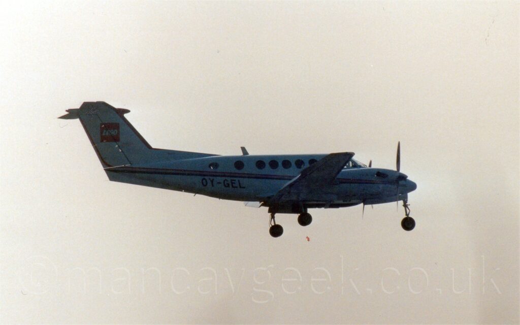 Slightly grainy and backlit side view of a white, twin propellor-engined BizProp flying from left to right at a low altitude, with undercarriage lowered and flaps extended behind the wings, suggesting it is about to land. There is a red and black stripe running along the body, as well as on the engine cowling, and the registration "OY-GEL" on the lower rear fuselage in black. There is a red square with a white "Lego" logo on the tail. Bright but hazy sky, with a slight orange tinge, fills the rest of the frame.