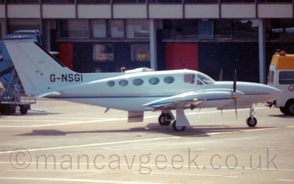 Side view of a white, twin engined BizProp parked facing to the right. The plane has a thin blue stripe running along the body, as well as along the sides of the engine cowlings. The registration "G-NSGI" is displayed prominently at the base of the tail in black. A door is open on the other side of the plane, visible hanging down below the body just behind the wing. A black and grey terminal building with red doors stretches across the top of the frame.