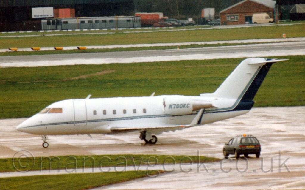 Slightly blurry side view of a white, twin engined BizJet taxiing from right to left. There is a thin blue stripe running along the body,. sweeping up to fill the rear of the tail. The same blue stripe fills the rear of the up-turned wingtips. The registration "N700KC" is on the side of the engine pods mounted on the sides of the rear fuselage. A grassed area and concrete apron fill the foreground, with more grass and a runway leading up to black hangars in the distance.