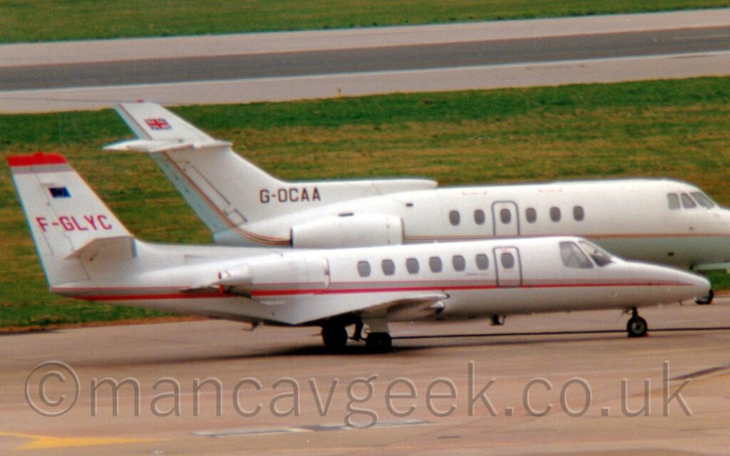 Side view of a pair of BizJets parked side by side, facing to the right. At the front is a long, thin, white BizJet with a red and black stripe running along the body and over the engine pods, which are mounted on the sides of the rear fuselage. The red registration "F-GLYC" is on the tail, under a similar stripe at the top of the tail Behind that is larger, slightly chunkier white BizJet with a very pale blue belly and thing golden stripe, both of which turn up under the white engine pods mounted on the sides of the rear fuselage, to fill the rear of the tail. The black registration "G-OCAA" is at the bottom of the tail, with a UK flag at the top. This plane's nose extends out of the frame to the right. Grey concrete apron fills the foreground, with wide patches of grass lining the sides of a runway in the background.