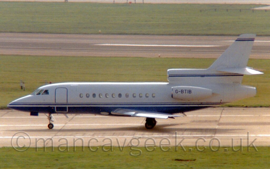 Side view of a white, 3 engined BizJet taxiing from right to left. The plane has a dark blue belly, with a dark blue stripe running along the body below the cabin windows, and along the top of the centre engine and tail. A large grassed area fills the foreground, with more grass separating a runway and another taxiway in the background.