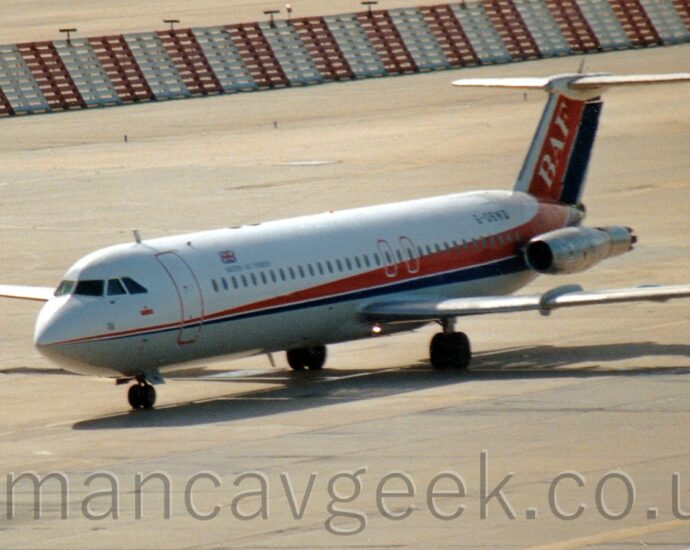 Side view of a white, twin engined jet airliner taxiing from right to left. The plane has a grey belly, and a thin red and blue stripe running along the body, getting wider as it moves rearwards, then sweeping up to fill the tail. The engines, mounted on the sides of the rear fuselage, are in a bare metal state. The registration "G-OBWD" is visible on the upper rear fuselage. Grey concrete taxiways fill almost all the frame, apart from a red and white banded metal barrier stretching across the top of the frame.