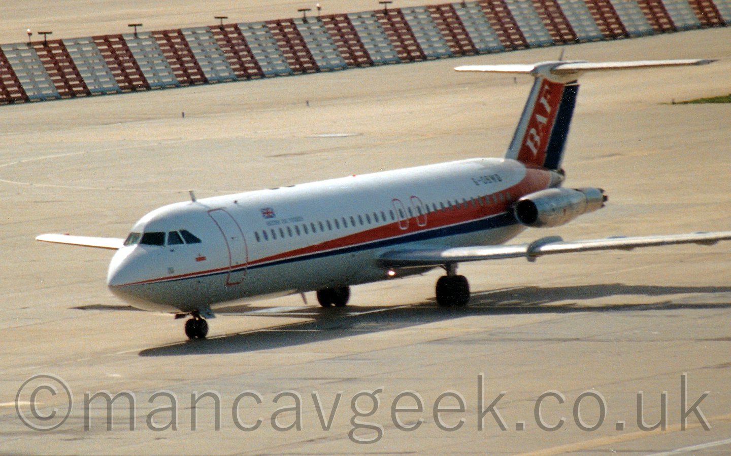 Side view of a white, twin engined jet airliner taxiing from right to left. The plane has a grey belly, and a thin red and blue stripe running along the body, getting wider as it moves rearwards, then sweeping up to fill the tail. The engines, mounted on the sides of the rear fuselage, are in a bare metal state. The registration "G-OBWD" is visible on the upper rear fuselage. Grey concrete taxiways fill almost all the frame, apart from a red and white banded metal barrier stretching across the top of the frame.