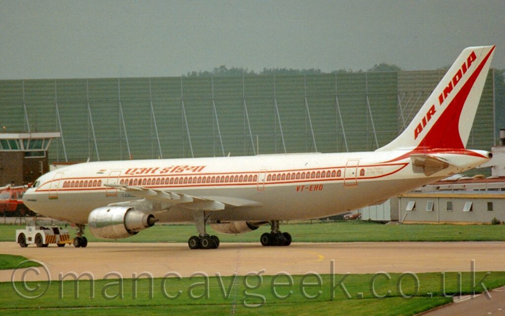 Side view of a twin engined jet airliner being towed from right to left along a grey taxiway by a white tug attached to the nosewheel. The plane has a white top half, with a grey lower half. A pair of thin red stripes runs along the body, above and below the passenger cabin windows, which themselves are outlined to resemble arched castle windows. There are red titles in a Sanskrit script on the upper forward fuselage, just aft of a small Indian flag, and the red registration "VT-EHO" on the lower rear. A stylised red flying bird, vaguely resembling a very pointy triangle, is on the white tail, with diagonal red "Air India" titles along the leading edge. Green grass lines the taxiway in the foreground, with a boxey, green metal frame filling the background, under grim grey sky.