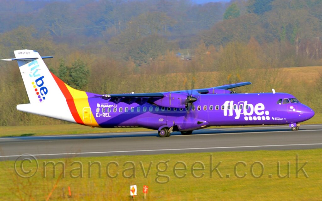 Side view of a high-winged, twin propellor-engined airliner taxiing from left to right off a black runway. he plane is mostly purple, with a white rear fuselage and tail, with large white "FlyBe" titles over a series of 6 white dots on the forward fuselage, repeated diagonally on the white tail in light and dark blue, and with coloured dots, 2 each of yellow, red, and purple. There are white "Stobart Air" titles on the upper rear fuselage, above the rear cabin windows, with the white registration "EI-REL" underneath the windows. Green grass fills the foreground, with trees and more grass stretching across the background, and more trees leading off into the distance.