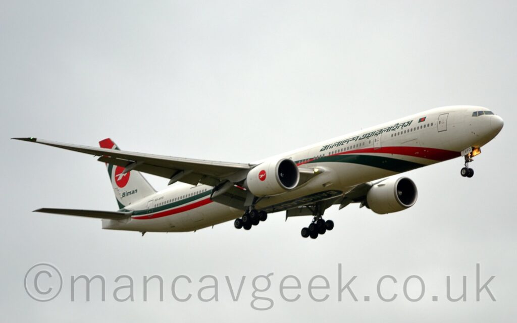 Side view of a twin engined jet airliner flying from left to right at a low altitude, with undercarriage lowered, and flaps extended from the rear of the wings, suggesting it is about to land. The plane is largely white, with a green and red stripe coming up from under the lower forward fuselage and running along the body. There are green titles, transliterating as "Biman Bangladesh Airlines" in a Sanskrit script on the upper forward fuselage, next to a small, green and red Bangladeshi flag. The large white engine pods under the wings have a red circle in the middle, containing a highly stylised white image of a flying bird. The same logo appears in a slightly larger form on the tail, with a curved green stripe running around it's rear, and a red stripe at the top. The black registration "S2-AFO" appears on the upper rear fuselage, the last 3 letters "AFO" also appearing on the open nose wheel doors, where a bright light is shining forwards. A green ovoid under the wingbox has the white word "Bangladesh", inverted from this perspective. Flat grey sky fills the rest of the frame.