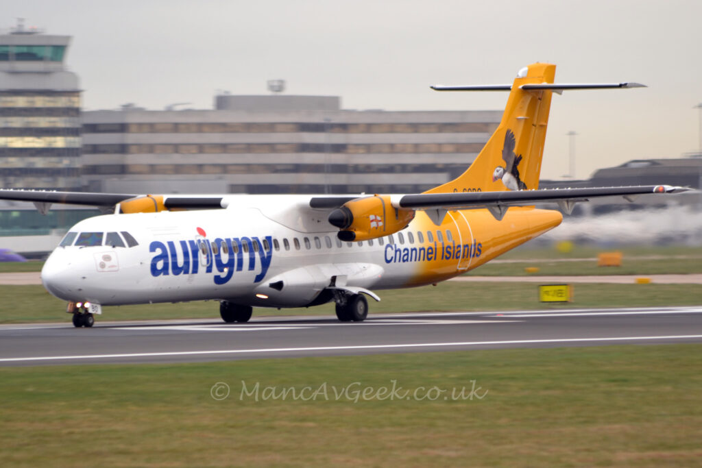 Side view of a high-winged, twin propellor-engined airliner moving at speed from right to left along a black runway. The plane is white at the front, with dark blue "Aurigny" titles on the side of the forward fuselage, while the rear is yellow, with blue "Channel Islands" text on the lower rear fuselage. The black registration "G-COBO" is on the bottom of the yellow tail, partially obscured by the wing. An image of a Puffin, a bird native to the Channel Islands, is on the tail. The yellow engine pods in front of the wings have a white flag with a red cross in the middle with a smaller yellow cross in the middle of that. Green grass fills the foreground, with more grass in the background on the other side of the runway leading up to a grey and black terminal building in the background. Grey sky fills the rest of the frame.