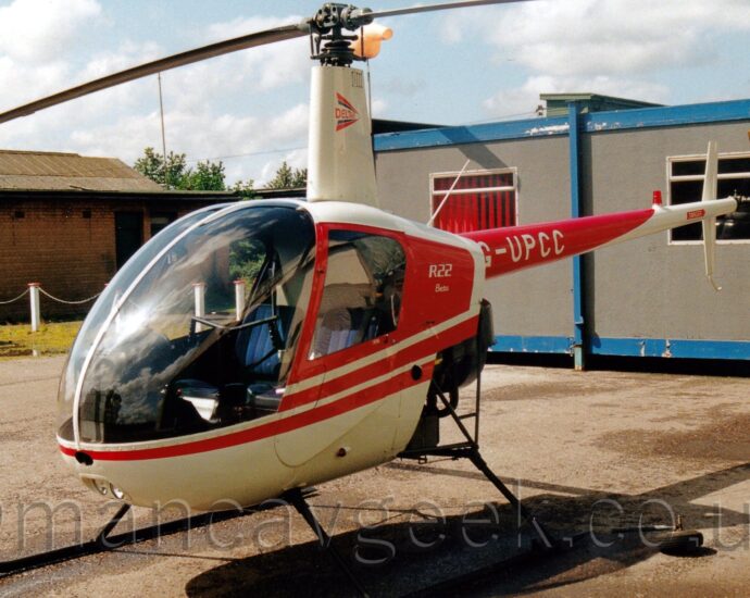 Side view of a light helicopter parked on grey concrete facing to the left. The chopper is mostly white, with red trim around the cockpit and on the tail boom. The tail boom has the white registration "G-UPCC" on the red section. A grey prefabricated building with a blue frame is in the background on the right, while a brown brick building with a grey car parked in front is on the left.