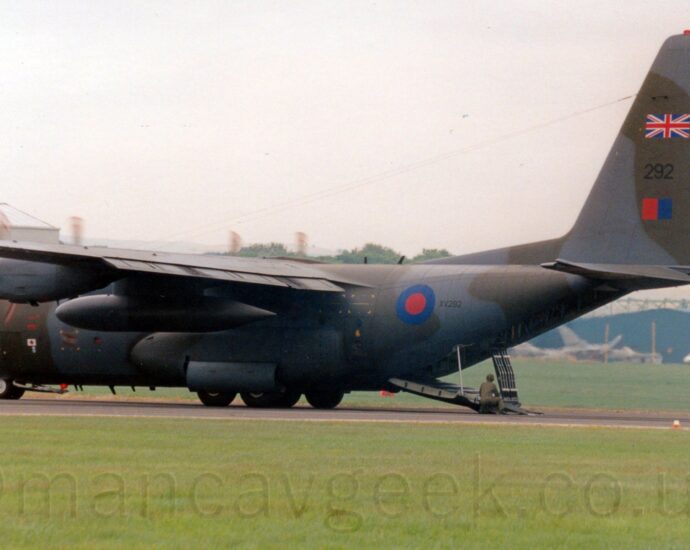 Side view of a high-winged, 4 propellor-engined military cargo plane parked on a black runway, with the red and white tips of the propellors a spinning blur indicating they are running full tilt, flaps extended from behind the wings, and the rear cargo loading ramp almost fully deployed, with a man wearing olive-green fatigues crouched by the end of the ramp. The plane is covered in an olive-green and grey camouflage scheme, with a blue and red roundel on the rear fuselage, next to the black serial number "VX232". There is a black number "232" on the nose under the cockpit, repeated on the tail, where it is sandwiched between a British flag above, and a red and blue fin flash below. Green grass fills the foreground, with more grass leading up to a tall grey hangar on the left, and a lower blue hangar on the left, with a couple of grey fighter jets in front of the latter. Grey sky fills the rest of the frame.