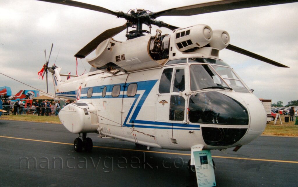 Side view of a white military transport helicopter parked facing to the left on a black tarmac taxiway. The helicopter has a blue stripe running along the body and tail boom, with a zig-zag down to below the cockpit windows. Sponsons on the rear fuselage house the main landing gear. A large panel that normally covers the engine has been dropped down, exposing the inner workings. A large sign has been placed against the nose, giving some detail about the aircraft. In the background, crowds of people are visible behind waist-level metal fences, with some tents and other exhibits behind them. Dismal grey skies fill the rest of the frame.