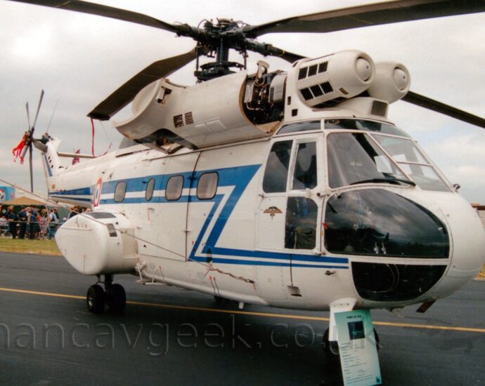 Side view of a white military transport helicopter parked facing to the left on a black tarmac taxiway. The helicopter has a blue stripe running along the body and tail boom, with a zig-zag down to below the cockpit windows. Sponsons on the rear fuselage house the main landing gear. A large panel that normally covers the engine has been dropped down, exposing the inner workings. A large sign has been placed against the nose, giving some detail about the aircraft. In the background, crowds of people are visible behind waist-level metal fences, with some tents and other exhibits behind them. Dismal grey skies fill the rest of the frame.