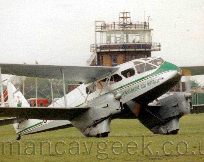Side view of a twin propellor-engined, bi-plane, pre-World War 2-era airliner taxiing from left to right on a grass airfield. The plane has a white top half with a grey belly, with a green and black stripe running along the body, and green "Caernarfon Air World" titles on the lower forward fuselage. A man can been seen sitting in the highly glazed cockpit, with a woman staring out from one of the curved, angular passenger cabin windows. A brown brick control tower can be seen in the middle of the background, with a couple of parked light aircraft on the left, and a red fire truck on the right, under flat grey sky.
