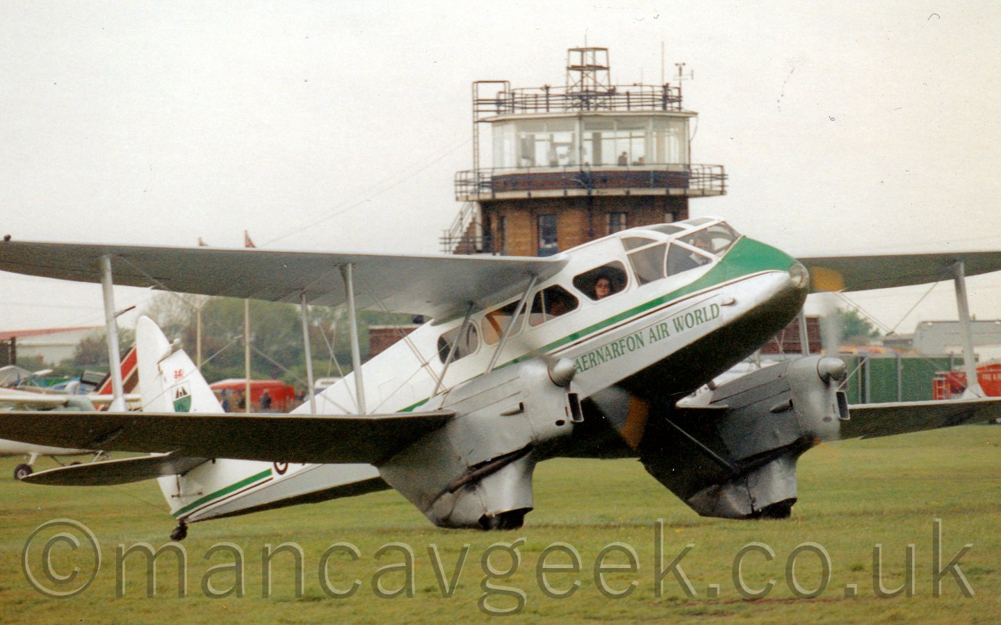 Side view of a twin propellor-engined, bi-plane, pre-World War 2-era airliner taxiing from left to right on a grass airfield. The plane has a white top half with a grey belly, with a green and black stripe running along the body, and green "Caernarfon Air World" titles on the lower forward fuselage. A man can been seen sitting in the highly glazed cockpit, with a woman staring out from one of the curved, angular passenger cabin windows. A brown brick control tower can be seen in the middle of the background, with a couple of parked light aircraft on the left, and a red fire truck on the right, under flat grey sky.