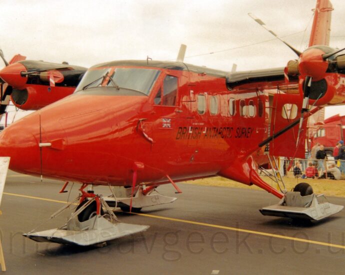 Front side view of a bright red, high-winged, twin propellor-engined parked facing to the left and slightly towards the camera. The plane's roof and the tops of the wings and engines are a shiny black, and black "British Antarctic Survey" are on the lower forward fuselage under the passenger cabin windows. A door in the rear fuselage is hanging open. Metal skids are mounted on the undercarriage, the wheels poking through the bottom. There is a white board on a wooden easel which has indecipherable text about this plane which partially obscures the nose on the left of the frame. Crowds of people behind a low metal fence are visible in the background, under flat white skies.