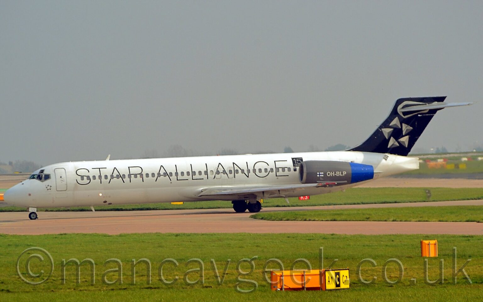 Side view of a white, twin engined jet airliner with the engines mounted on the sides of the rear fuselage, taxiing from right to left. The body is all white, with black "Star Alliance" titles running the whole length. The tail is black, with a 5-pointed star made up of grey pyramids, arranged in a circle. The engine pods are grey with blue exhausts, with the registration "OH-BLP" in white, next to a Finnish flag. Green grass surrounding taxiways fill the foreground, with more grass and a runway in the background, under grey-blue skies.