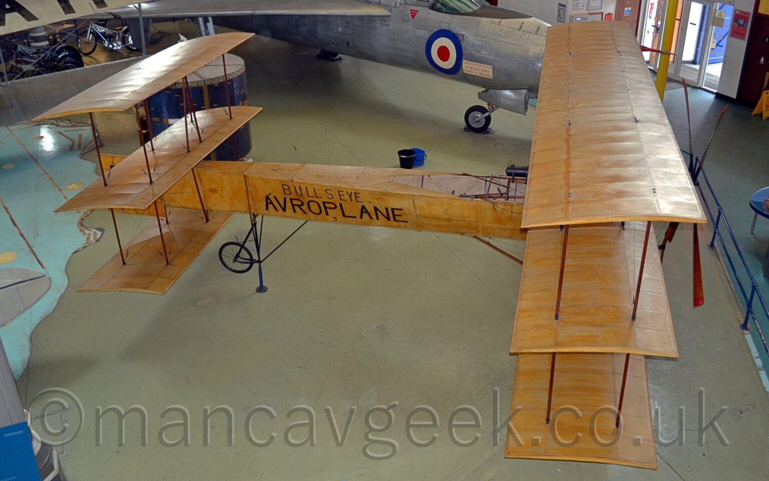 Elevated side view of a single-engined tri-plane on display facing to the right in a museum. The doped-cloth skin of the plane has a streaky, light brown colour, similar to varnished oak. As well as having 3 stacked wings at the front, there are 3 horizontal stabilisers at the rear. The black words "Bullseye" and "Avroplane" are visible on the sides of the rear fuselage. Creamy-grey floor fills moist of the foreground and part of the background, with a military jet fighter plane visible at the top of the frame.