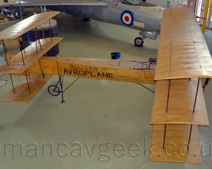 Elevated side view of a single-engined tri-plane on display facing to the right in a museum. The doped-cloth skin of the plane has a streaky, light brown colour, similar to varnished oak. As well as having 3 stacked wings at the front, there are 3 horizontal stabilisers at the rear. The black words "Bullseye" and "Avroplane" are visible on the sides of the rear fuselage. Creamy-grey floor fills moist of the foreground and part of the background, with a military jet fighter plane visible at the top of the frame.