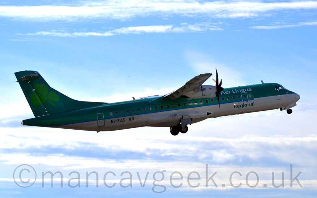 Side view of a green, high-winged, twin propellor-engined airliner flying from left to right at a low altitude, with flaps extended from the rear of the wings, undercarriage retracting, and the nose raised sharply, suggesting it has just taken off. The planes top half is a dark green, with a lighter green stripe over the passenger cabin windows, and a white belly. There are white "Aer Lingus" titles on the upper forward fuselage, and green "Regional" titles at the bottom. The black registration "EI-FAS" is on the lower rear fuselage, next to a small Irish flag. There is a large, light green shamrock leaf on the tail. Pale blue sky with streaks of white cloud fill the rest of the frame.