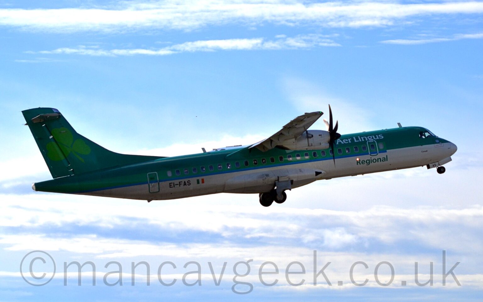 Side view of a green, high-winged, twin propellor-engined airliner flying from left to right at a low altitude, with flaps extended from the rear of the wings, undercarriage retracting, and the nose raised sharply, suggesting it has just taken off. The planes top half is a dark green, with a lighter green stripe over the passenger cabin windows, and a white belly. There are white "Aer Lingus" titles on the upper forward fuselage, and green "Regional" titles at the bottom. The black registration "EI-FAS" is on the lower rear fuselage, next to a small Irish flag. There is a large, light green shamrock leaf on the tail. Pale blue sky with streaks of white cloud fill the rest of the frame.