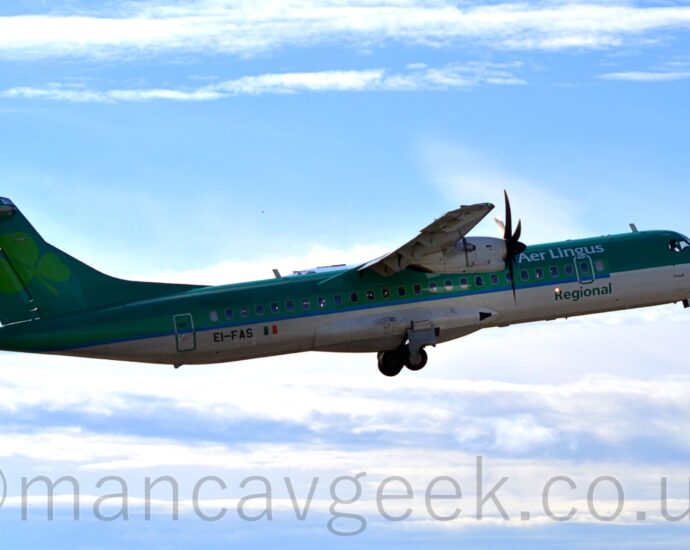 Side view of a green, high-winged, twin propellor-engined airliner flying from left to right at a low altitude, with flaps extended from the rear of the wings, undercarriage retracting, and the nose raised sharply, suggesting it has just taken off. The planes top half is a dark green, with a lighter green stripe over the passenger cabin windows, and a white belly. There are white "Aer Lingus" titles on the upper forward fuselage, and green "Regional" titles at the bottom. The black registration "EI-FAS" is on the lower rear fuselage, next to a small Irish flag. There is a large, light green shamrock leaf on the tail. Pale blue sky with streaks of white cloud fill the rest of the frame.