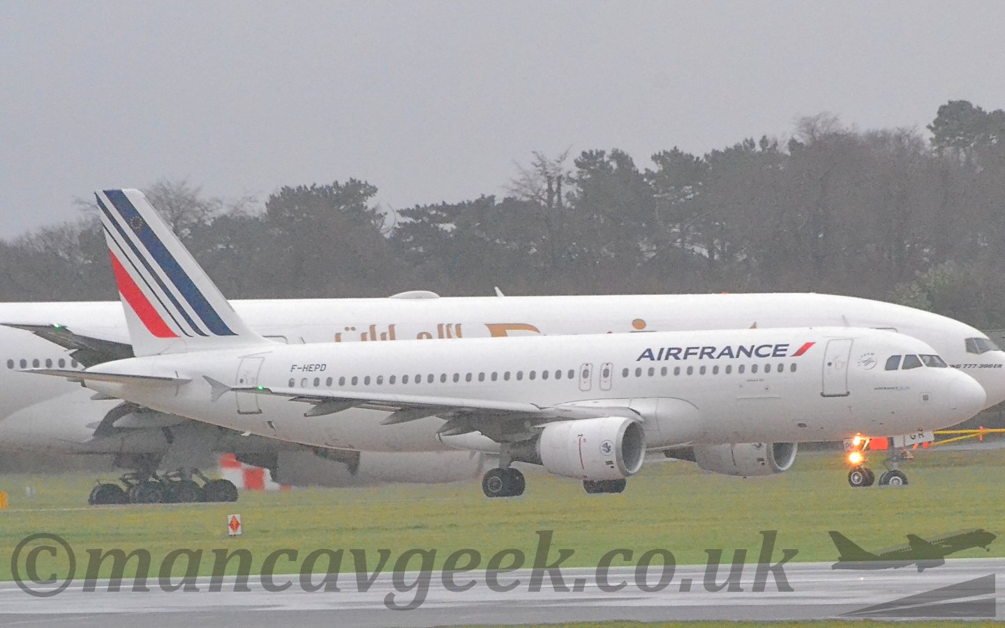 Side view of a twin engined jet airliner taxiing from left to right. The plane is mostly white, with blue "Air France" titles on the upper forward fuselage, the black registration "F-HEPD" on the upper rear, and 3 diagonal blue stripes on the tail, getting narrower as they move rearwards, followed by a single red stripe. There is a single bright light on the nose-wheel, flaring in the camera lens. A black runway and a wide grassed area fill the foreground, with a larger, white, twin-engined jet airliner in the background. Trees beyond that meet flat grey sky.