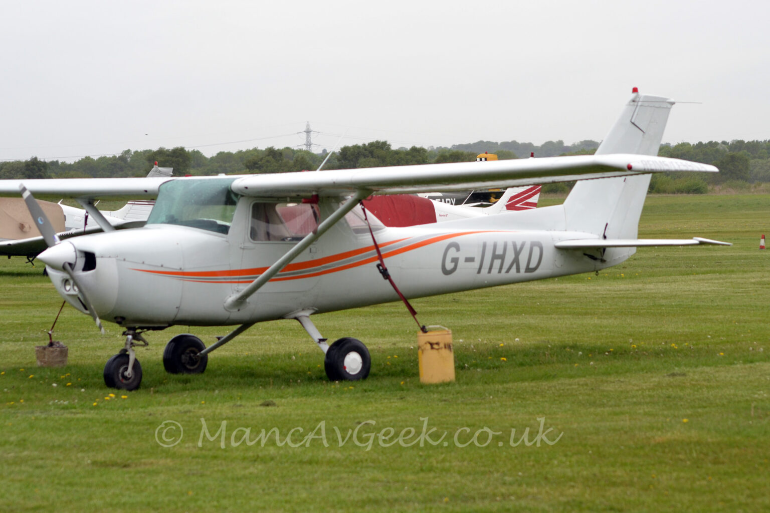 Side view of a high winged, single engined light aircraft parked facing to the left on grass, with large weights attached to the wings by long red straps to keep it in place. The plane is mostly white, with an orange and grey swirl reaching from near the front of the engine cowling, below and the up around the back of the cockpit, and the silver registration "G-IHXD" on the side sf the rear fuselage. Green grass fills the foreground and a large part of the background, with a couple more planes parked on the left of the frame behind this one, and trees in the distance, meeting flat grey sky.