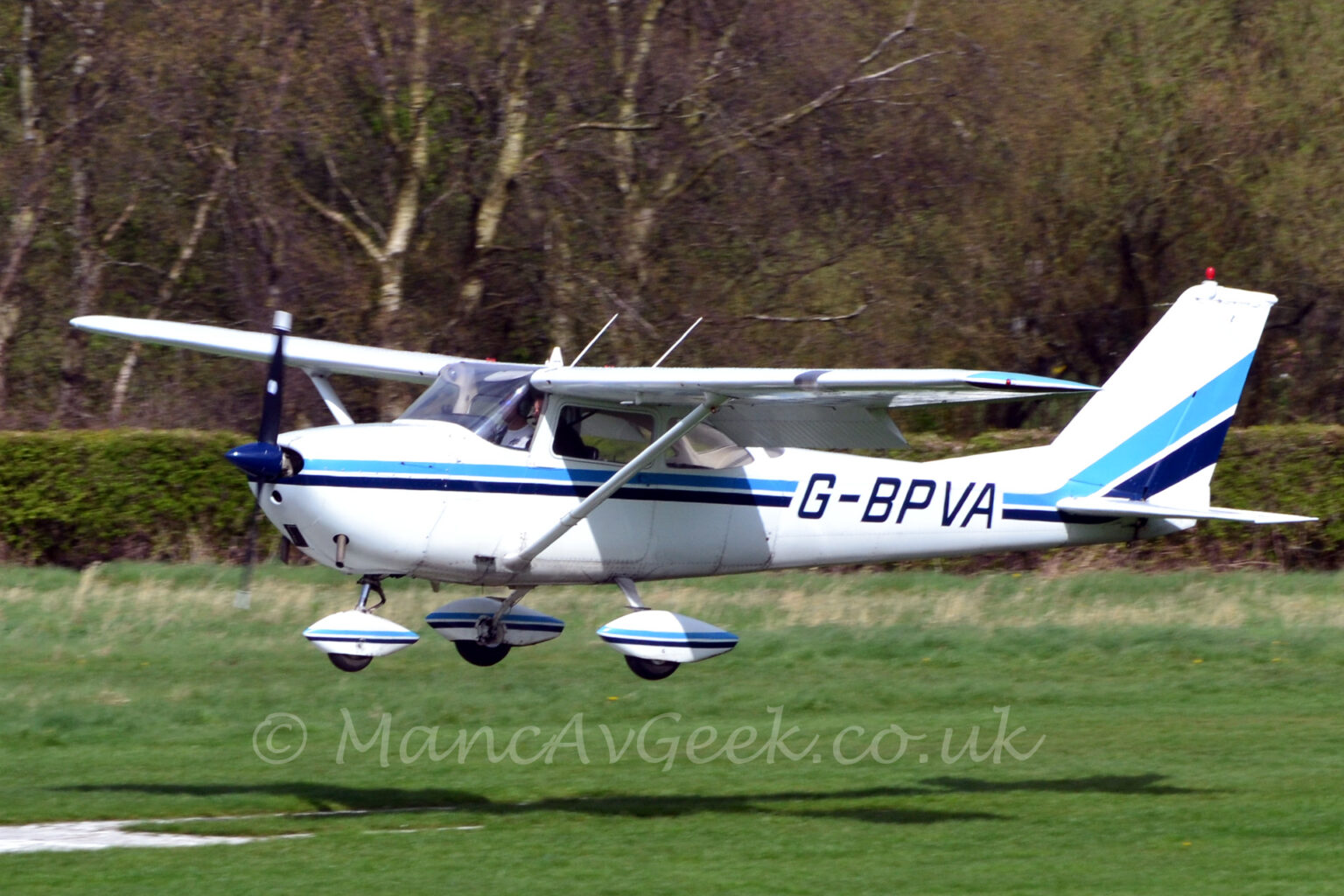 Side view of a high-winged, single-engined light aircraft flying from right to left at a very low altitude, just a couple of metres above the white numbers on a grass runway. The plane is mostly white, with a light and dark blue stripe running along the body and up into the lower parts of the tail, overlaid with dark blue registration "G-BPVA" on the rear fuselage. The plane has flaps extended from the rear of the wings, and is flying more or less straight and level, suggesting it is just about to land. Green grass fills the foreground, with trees filling the background.