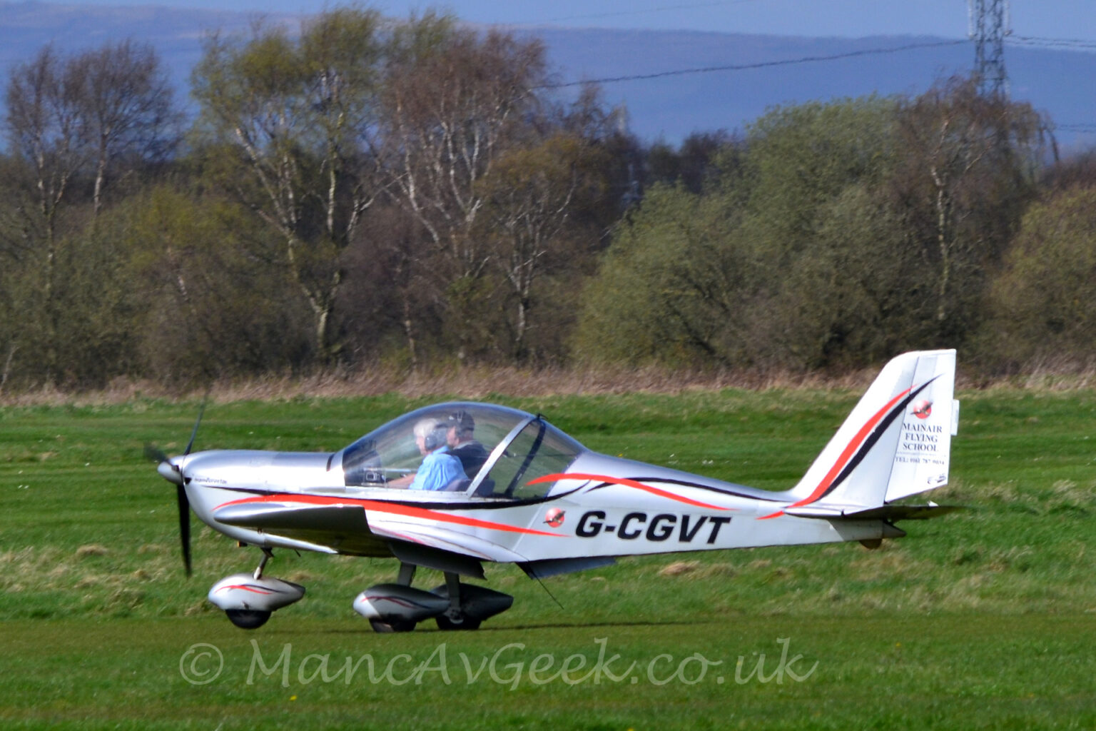 Side view of a single engined light aircraft taxing from right to left across a grass airfield. The plane is a silvery grey, with a black and red stripe swirling it's way along the body, and the black registration "G-CGVT" on the lower rear fuselage. There is black text "Mainair Flying School", alongside a telephone number on the rear half of the tail. Metallic grey spats with black and red stripes cover the wheels on the undercarriage. Two people can be seen in the cockpit through the comically oversized bubble canopy. Green grass fills the foreground and most of the background, with trees on the airfield perimeter, rolling hills in the far distance, and blue sky at the top of the frame.