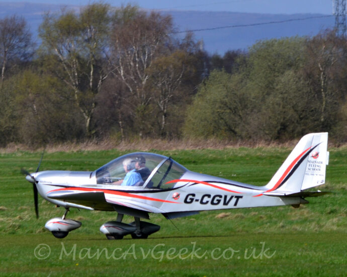 Side view of a single engined light aircraft taxing from right to left across a grass airfield. The plane is a silvery grey, with a black and red stripe swirling it's way along the body, and the black registration "G-CGVT" on the lower rear fuselage. There is black text "Mainair Flying School", alongside a telephone number on the rear half of the tail. Metallic grey spats with black and red stripes cover the wheels on the undercarriage. Two people can be seen in the cockpit through the comically oversized bubble canopy. Green grass fills the foreground and most of the background, with trees on the airfield perimeter, rolling hills in the far distance, and blue sky at the top of the frame.
