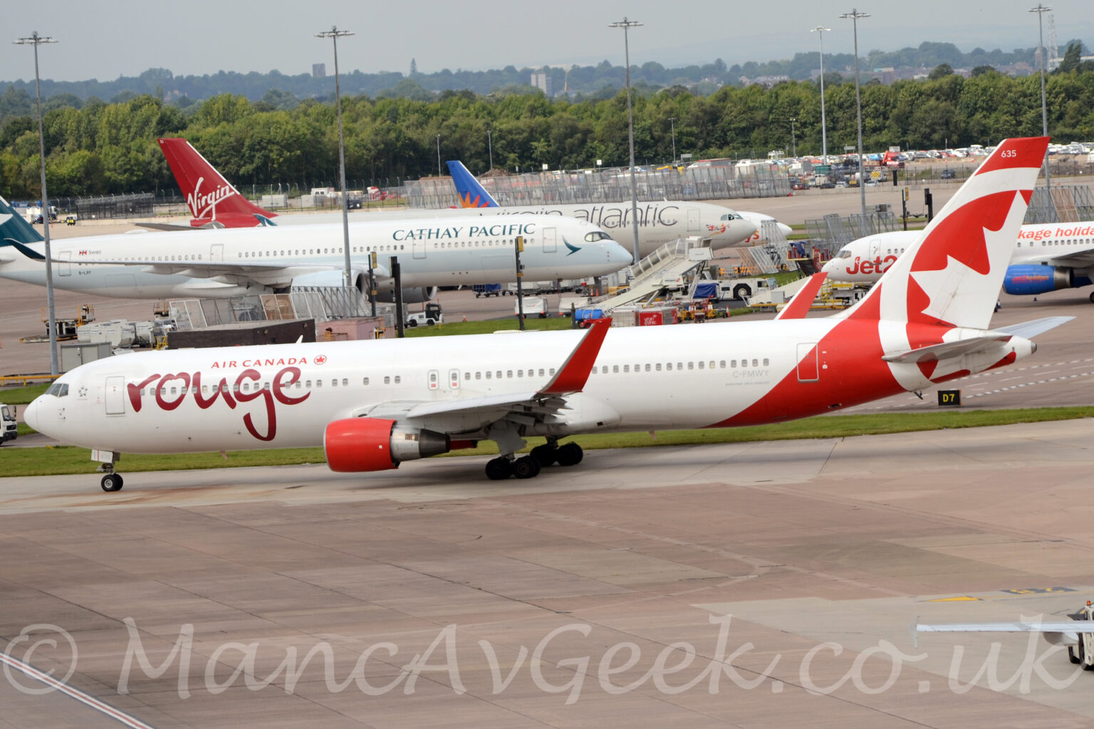 Side view of a twin engined jet airliner taxiing from right to left along a grey taxiway. The plane is largely white, with red "Air Canada" titles on the upper forward fuselage, next to a red maple leaf in the red outline of a circle, with larger, seemingly hand-written "Rouge" text in a deeper red below. The pale grey registration "C-FMWY" is on the lower rear fuselage, below the rear-most windows. The tail and rear fuselage are red, with part of a larger, white version of the circle and maple leaf logo, and the white number "635" right at the top. The engine pods under the wings are bright red, as are the tall, up-turned wing-tips. Grey concrete apron fills the foreground, with several more plane parked on more apron in the background, behind a row of tall lighting poles reaching into the grey sky. A ground level car park is beyond these other planes, with trees beyond that.
