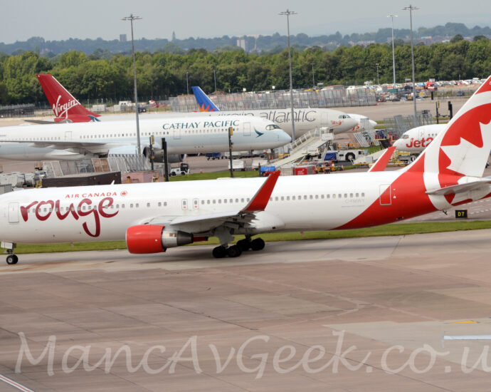 Side view of a twin engined jet airliner taxiing from right to left along a grey taxiway. The plane is largely white, with red "Air Canada" titles on the upper forward fuselage, next to a red maple leaf in the red outline of a circle, with larger, seemingly hand-written "Rouge" text in a deeper red below. The pale grey registration "C-FMWY" is on the lower rear fuselage, below the rear-most windows. The tail and rear fuselage are red, with part of a larger, white version of the circle and maple leaf logo, and the white number "635" right at the top. The engine pods under the wings are bright red, as are the tall, up-turned wing-tips. Grey concrete apron fills the foreground, with several more plane parked on more apron in the background, behind a row of tall lighting poles reaching into the grey sky. A ground level car park is beyond these other planes, with trees beyond that.