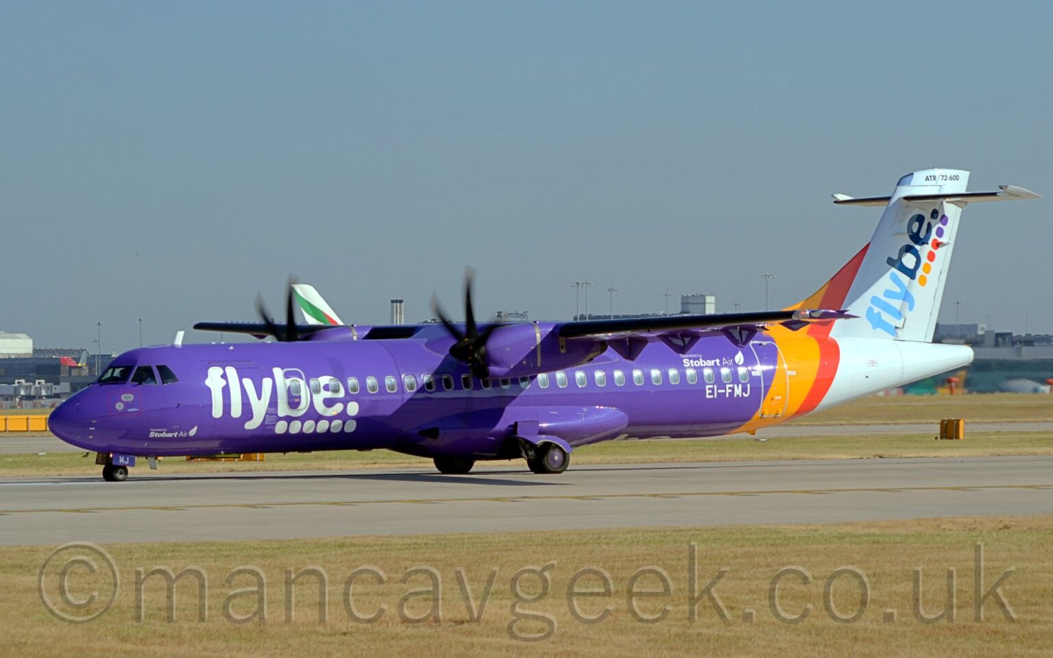 Side view of a high-winged, twin propellor-engined airliner taxiing from right to left along a grey runway. The plane is mostly purple, with large white "FlyBe" titles over 6 white dots., with much smaller, white "Stobart Air" titles next to a white feather on the lower forward fuselage, under the nose. Larger "Operated by Stobart Air" titles are over the rear cabin windows, just forward of the rear door, , with the white registration "EI-FMJ" just under those windows. Yellow and red diagonal bands around the rear body lead into the white tail and tail-cone, with diagonal two-tone blue "FlyBe" titles over 2 yellow, 2red, and 2 purple dots on the tail. At the top of the tail, just above the horizontal stabiliser, there is black "ATR 72-600" text. Green grass stretches across the foreground, lining the side of the runway,. In the background, more grass leads up to a grey terminal building in the distance, with the tail of a very large, white airliner visible over the top of this plane. Blue sky fills the rest of the frame.