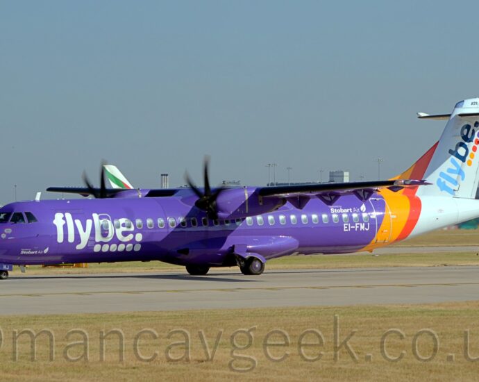 Side view of a high-winged, twin propellor-engined airliner taxiing from right to left along a grey runway. The plane is mostly purple, with large white "FlyBe" titles over 6 white dots., with much smaller, white "Stobart Air" titles next to a white feather on the lower forward fuselage, under the nose. Larger "Operated by Stobart Air" titles are over the rear cabin windows, just forward of the rear door, , with the white registration "EI-FMJ" just under those windows. Yellow and red diagonal bands around the rear body lead into the white tail and tail-cone, with diagonal two-tone blue "FlyBe" titles over 2 yellow, 2red, and 2 purple dots on the tail. At the top of the tail, just above the horizontal stabiliser, there is black "ATR 72-600" text. Green grass stretches across the foreground, lining the side of the runway,. In the background, more grass leads up to a grey terminal building in the distance, with the tail of a very large, white airliner visible over the top of this plane. Blue sky fills the rest of the frame.
