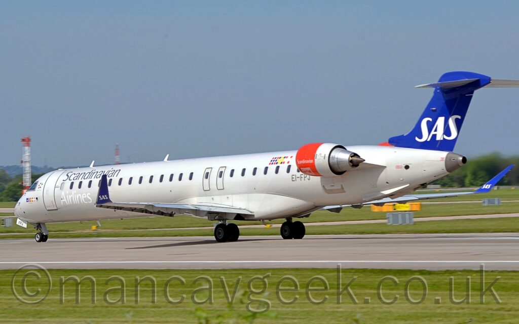 Side view of a long, thin, white, twin engined jet airliner with the engines mounted on the sides of the rear fuselage. The plane is moving from right to left and slightly away from the camera on a grey runway. The plane is almost entirely white, with silver "Scandinavian" titles on the upper forward fuselage, just above the first few passenger cabin windows, with even whiter "Airlines" titles on the lower forward fuselage, under the windows. The registration "EI-FPJ" is on the lower rear fuselage, under the last of the windows, just in front of the red engine pods. The tail is a deep blue, with white "SAS" titles. The engine pods are red at the front, with small white "Scandinavian" titles. The upturned wingtips are the same blue as the tail, with the same "SAS" logo. Green grass fills the foreground, with the odd stray branch of small trees at the bottom. More grass leads off to trees lining the airfield perimeter in the distance, under grey-blue skies.